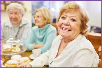 Three older adults sitting at a table with pastries and coffee, smiling and enjoying each other's company in a cozy indoor setting.