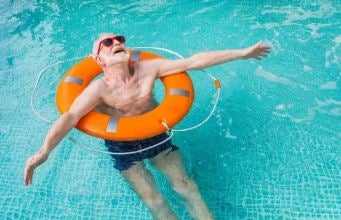 An elderly man wearing red sunglasses floats in a pool with an orange life ring around his chest, arms outstretched.