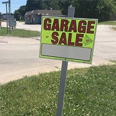 A "Garage Sale" sign with blank spaces for details is posted on a grassy area beside a street, with buildings visible in the background.