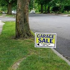 A "Garage Sale" sign with an arrow is placed on a grassy lawn next to a tree by the sidewalk, pointing toward a residential street.