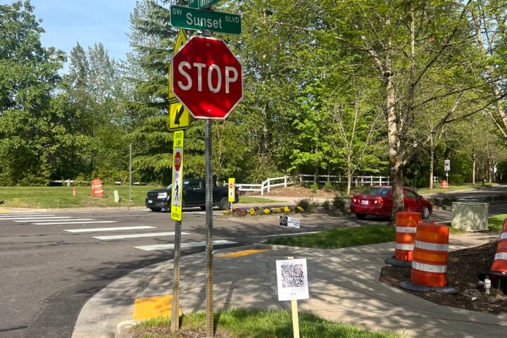 A stop sign at the corner of SW Sunset Blvd and Trimble with a QR code sign on the grass, orange traffic barrels, and parked cars in the background.