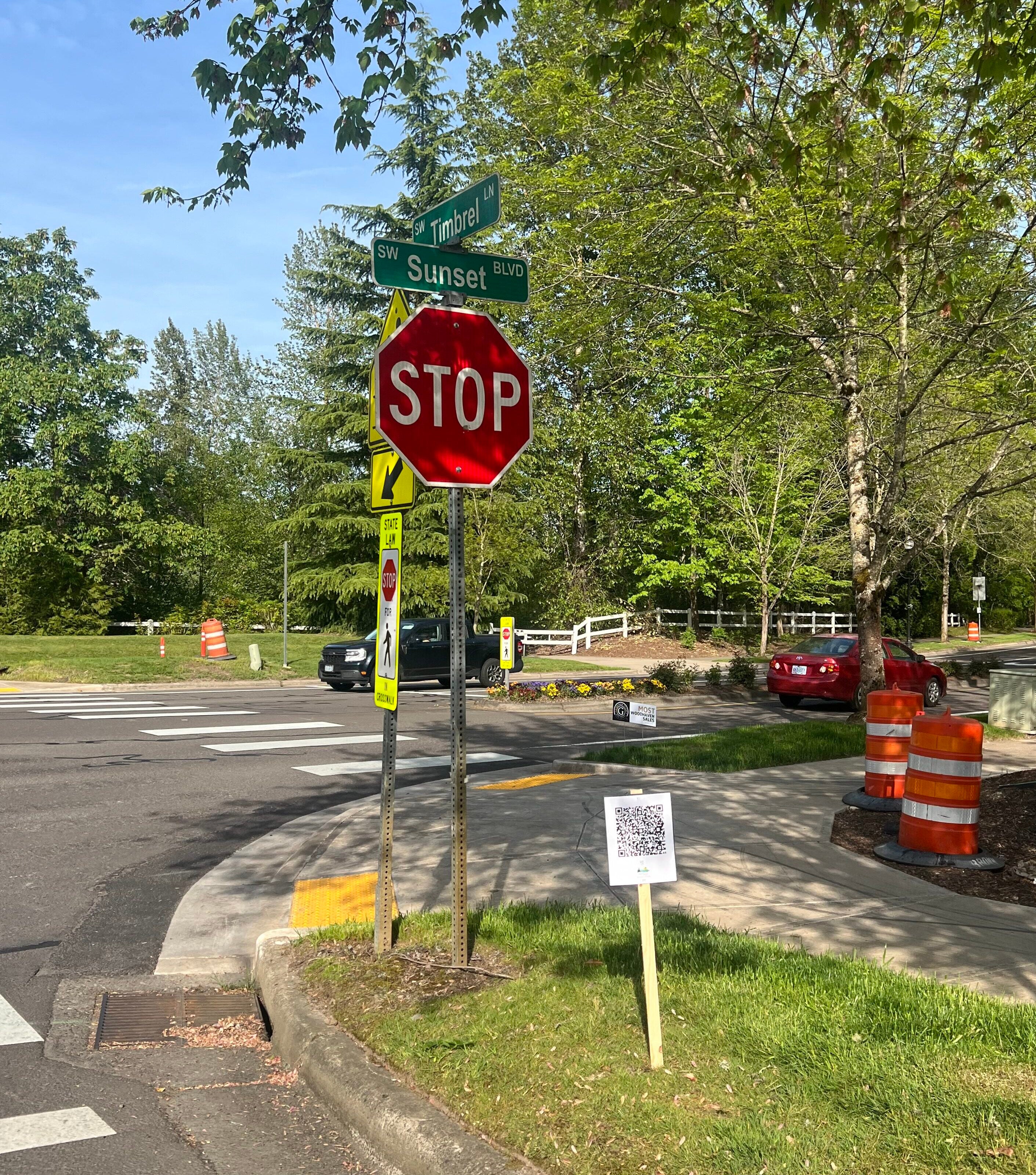 A stop sign at the corner of SW Sunset Blvd and Trimble with a QR code sign on the grass, orange traffic barrels, and parked cars in the background.