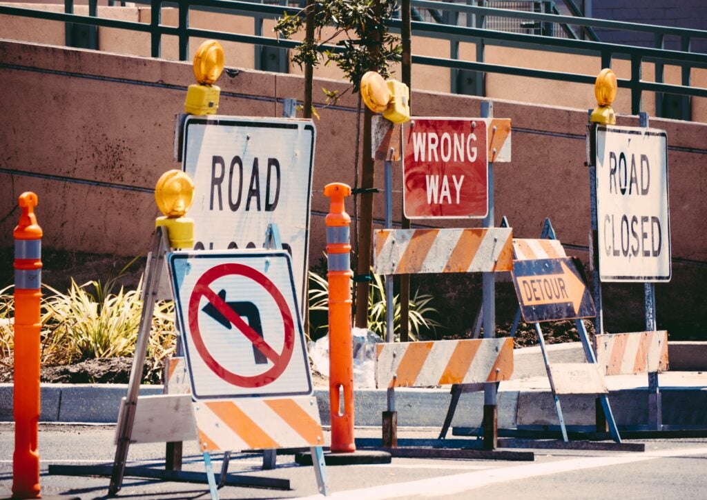 Multiple road signs and barricades, including "Road Closed," "Wrong Way," "No Left Turn," and "Detour," block a street, indicating construction or restricted access.