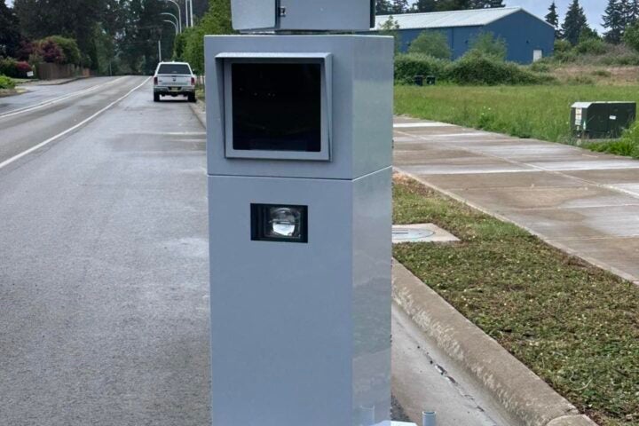 A speed camera installation on the side of a suburban road, with a vehicle and greenery visible in the background.