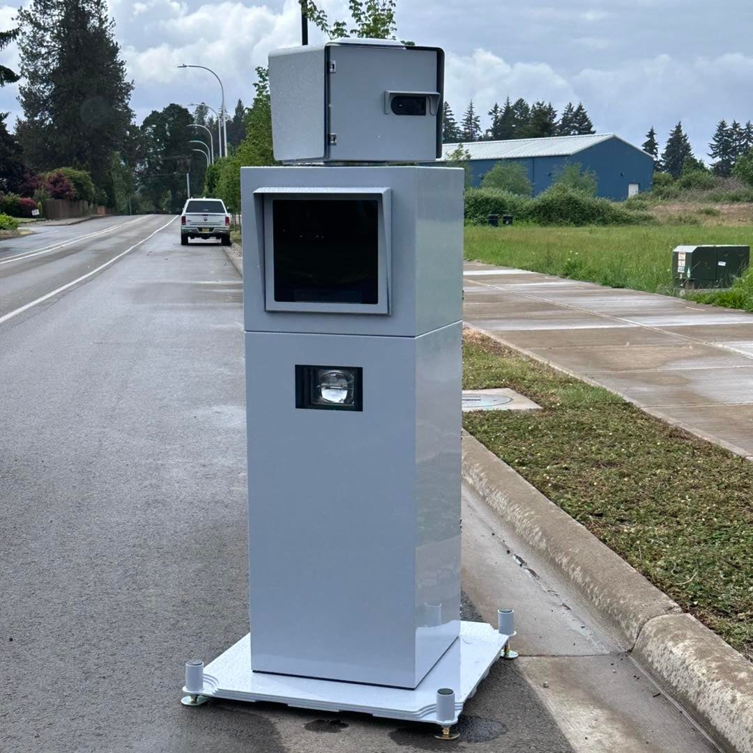 A speed camera installation on the side of a suburban road, with a vehicle and greenery visible in the background.