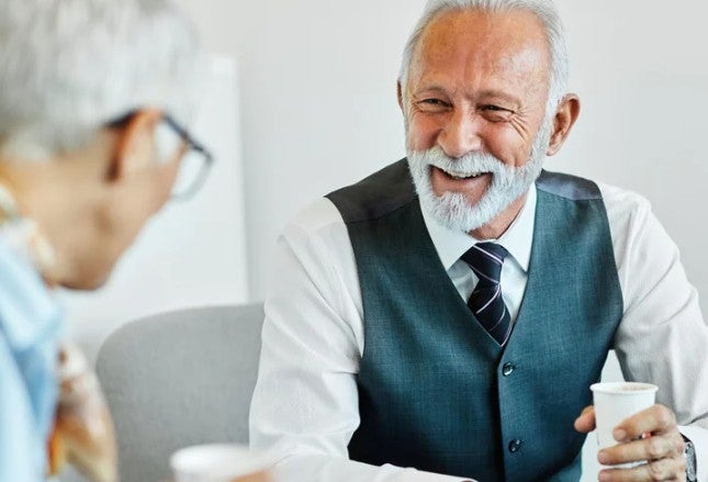 Two older men sit indoors, smiling and talking while holding disposable cups. One wears a vest and tie, the other has glasses.