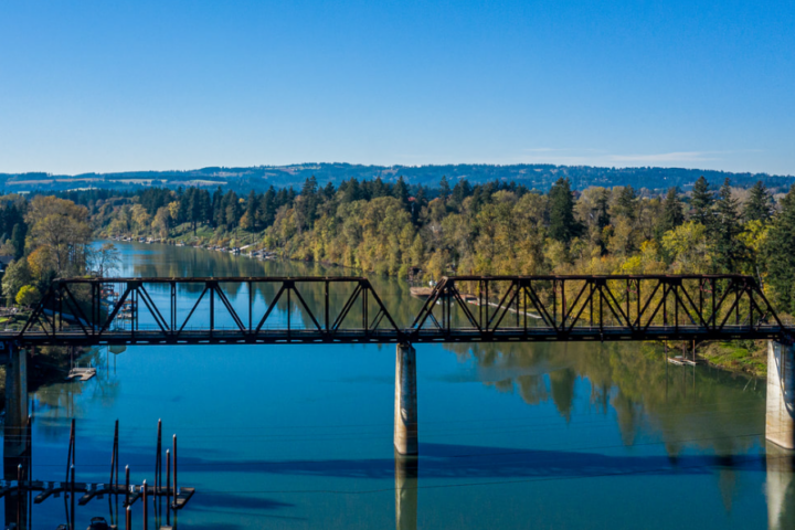 A steel truss railroad bridge crosses a calm river surrounded by trees and hills under a clear blue sky.
