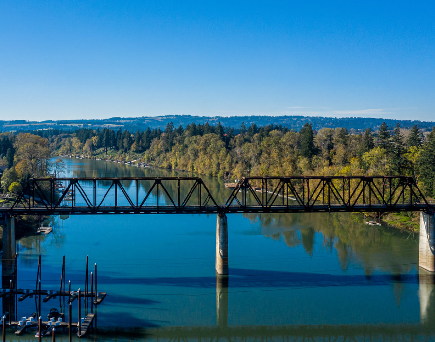A steel truss railroad bridge crosses a calm river surrounded by trees and hills under a clear blue sky.