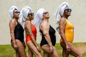 Four women in swimsuits and sunglasses stand in a line outdoors, each with a white towel wrapped around their head, facing to the right.