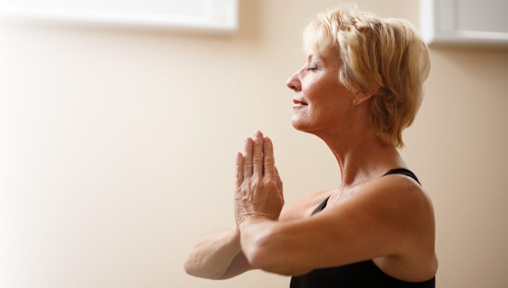 Older woman with short blonde hair wearing a black tank top practices yoga indoors, holding hands in prayer position and appearing calm and focused.