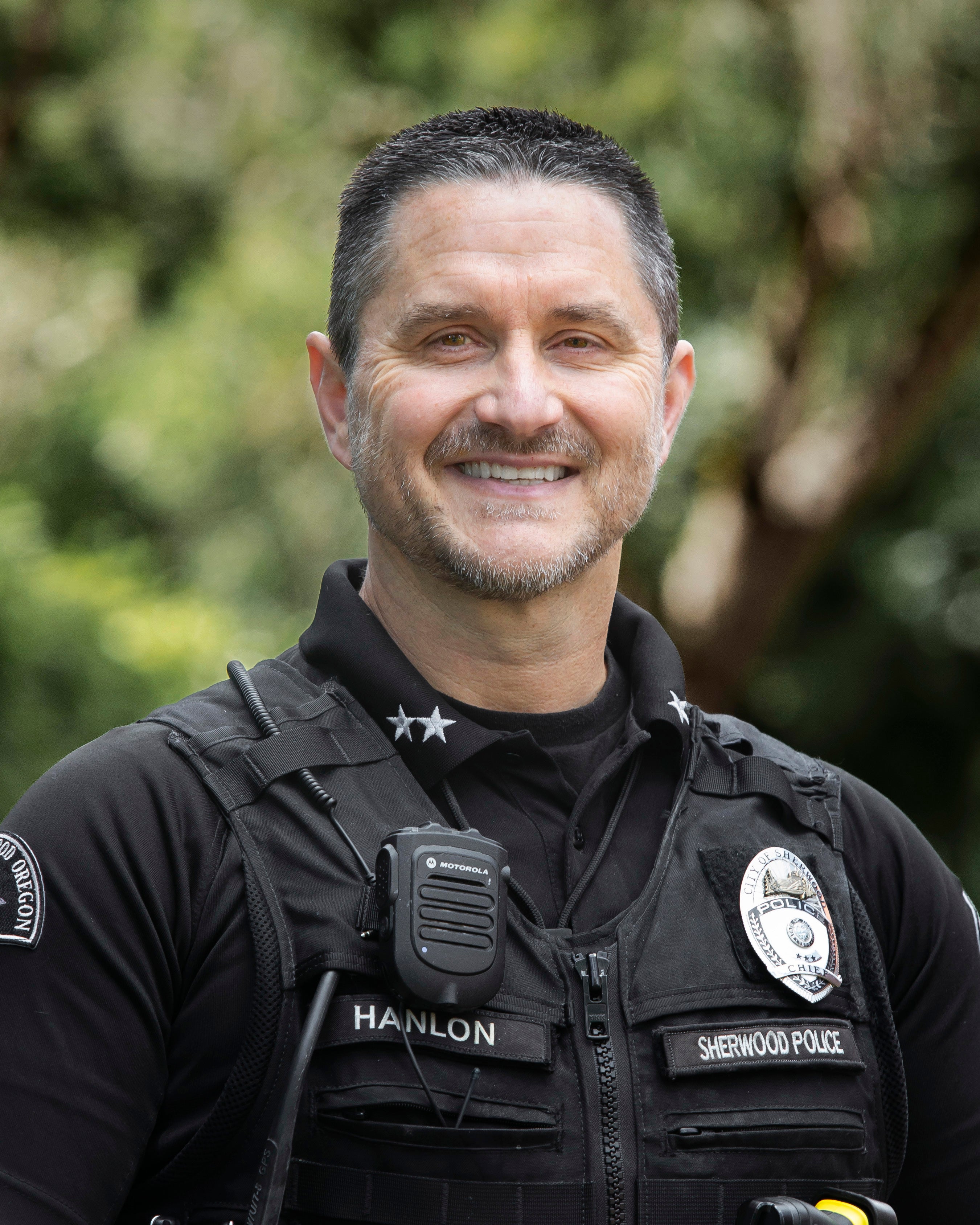 A police officer in uniform, wearing a vest labeled "Hanlon" and "Sherwood Police," smiles outdoors with trees in the background.