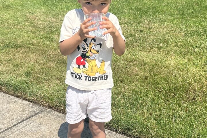 A young boy stands on a sidewalk drinking from a plastic cup, wearing a Mickey Mouse shirt, shorts, and sneakers, with grass and shrubs in the background.