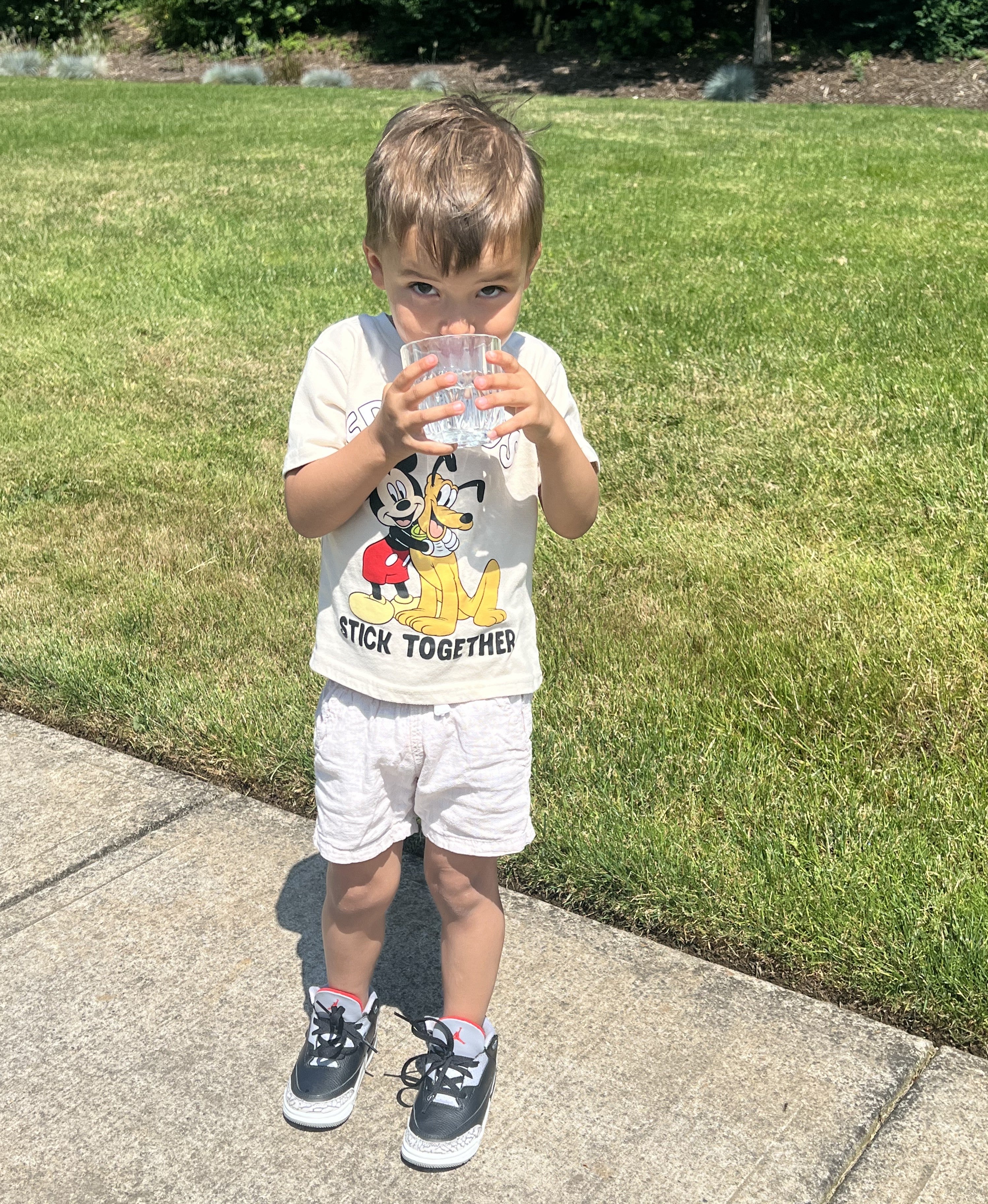 A young boy stands on a sidewalk drinking from a plastic cup, wearing a Mickey Mouse shirt, shorts, and sneakers, with grass and shrubs in the background.