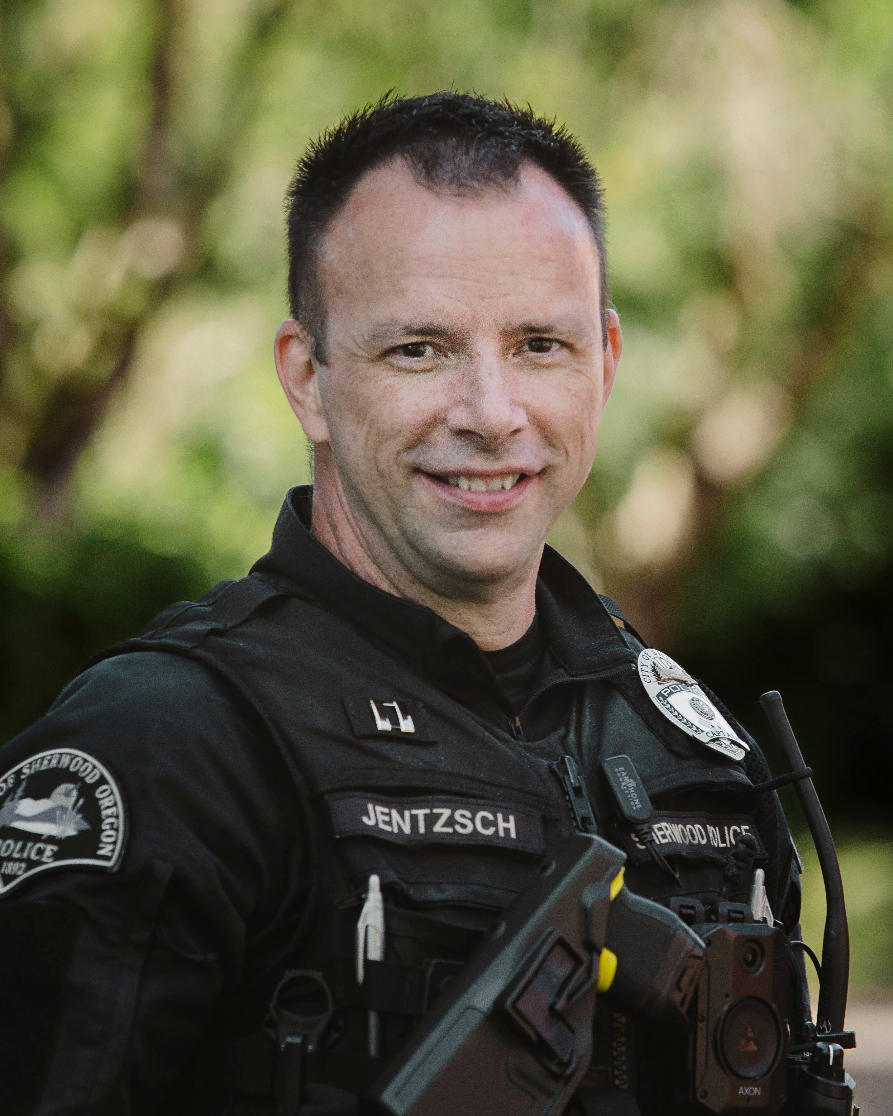A police officer in uniform stands outdoors, smiling at the camera. The name badge reads "Jentzsch" and he is wearing a body camera and communication equipment.