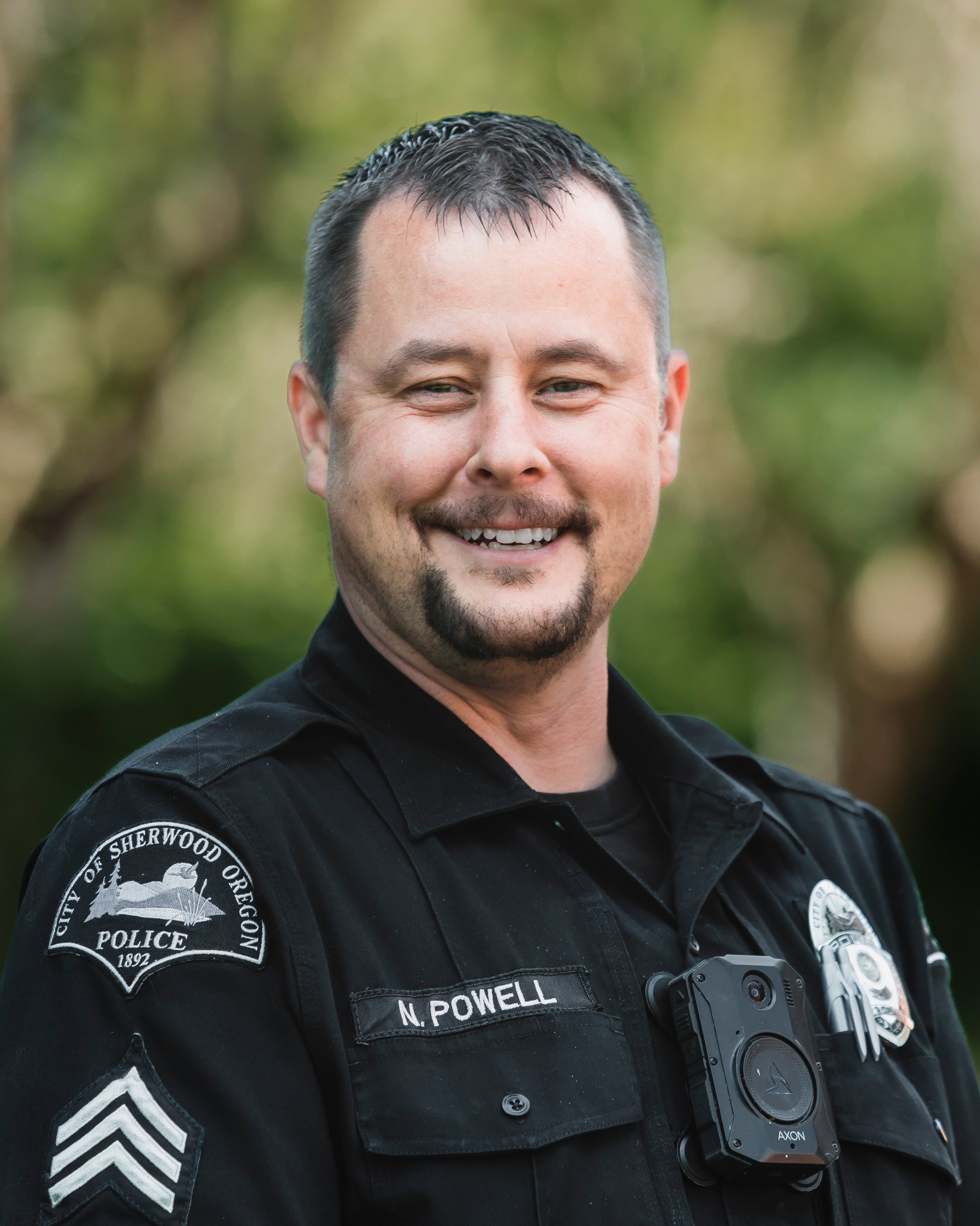 A police officer in uniform with a body camera, name tag "N. Powell," and a "City of Sherwood, Oregon Police" patch, standing outdoors.