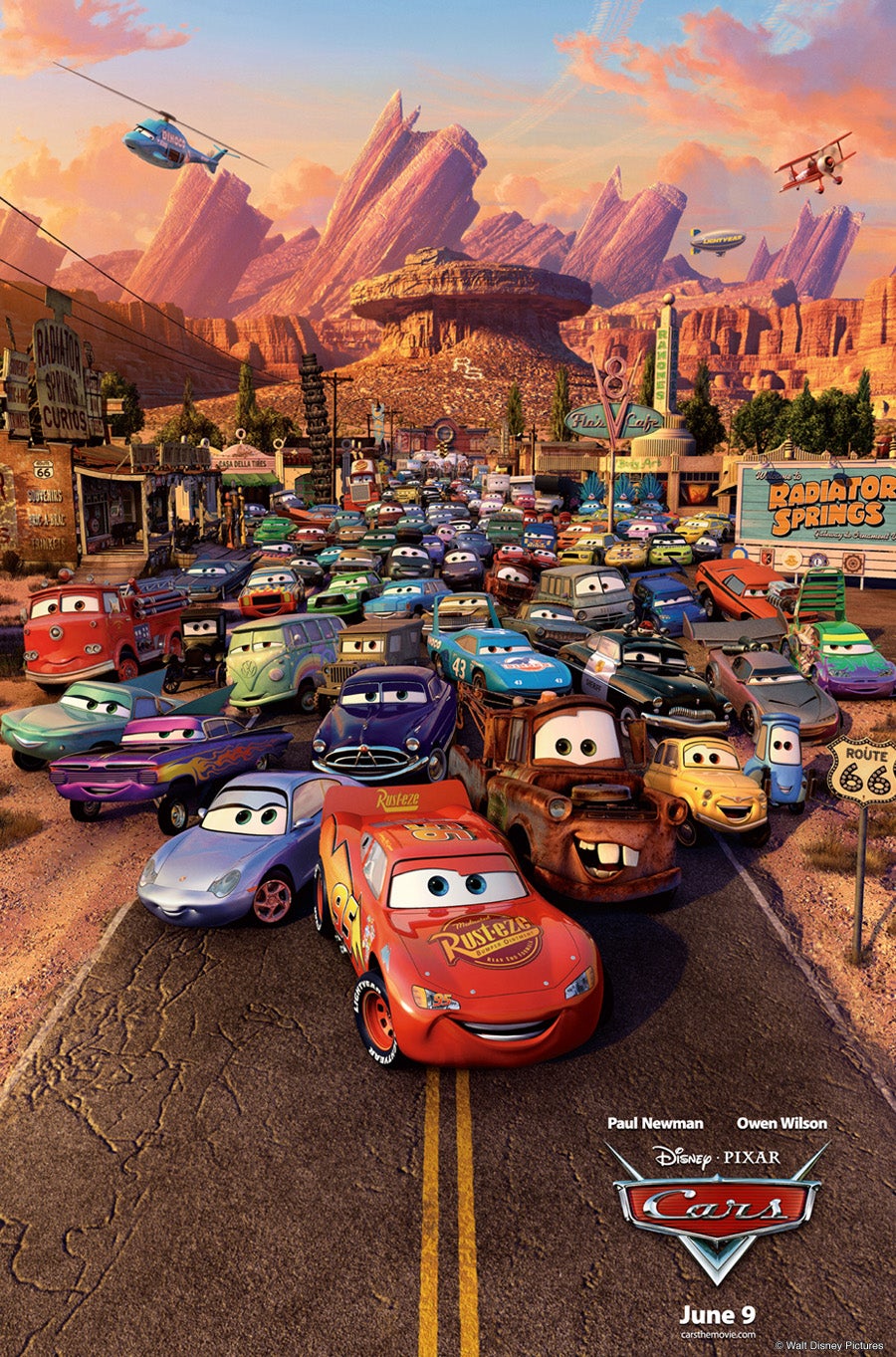Animated cars with expressive faces gather on a street in a desert town, with rock formations and a Radiator Springs sign in the background. Movie title "Cars" appears at the bottom.