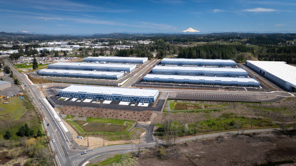 Aerial view of a large industrial complex with multiple warehouse buildings, parking lots, and nearby roads, surrounded by greenery and distant mountains under a blue sky.
