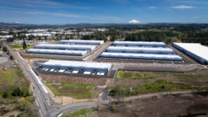 Aerial view of a large industrial complex with multiple warehouse buildings, parking lots, and nearby roads, surrounded by greenery and distant mountains under a blue sky.