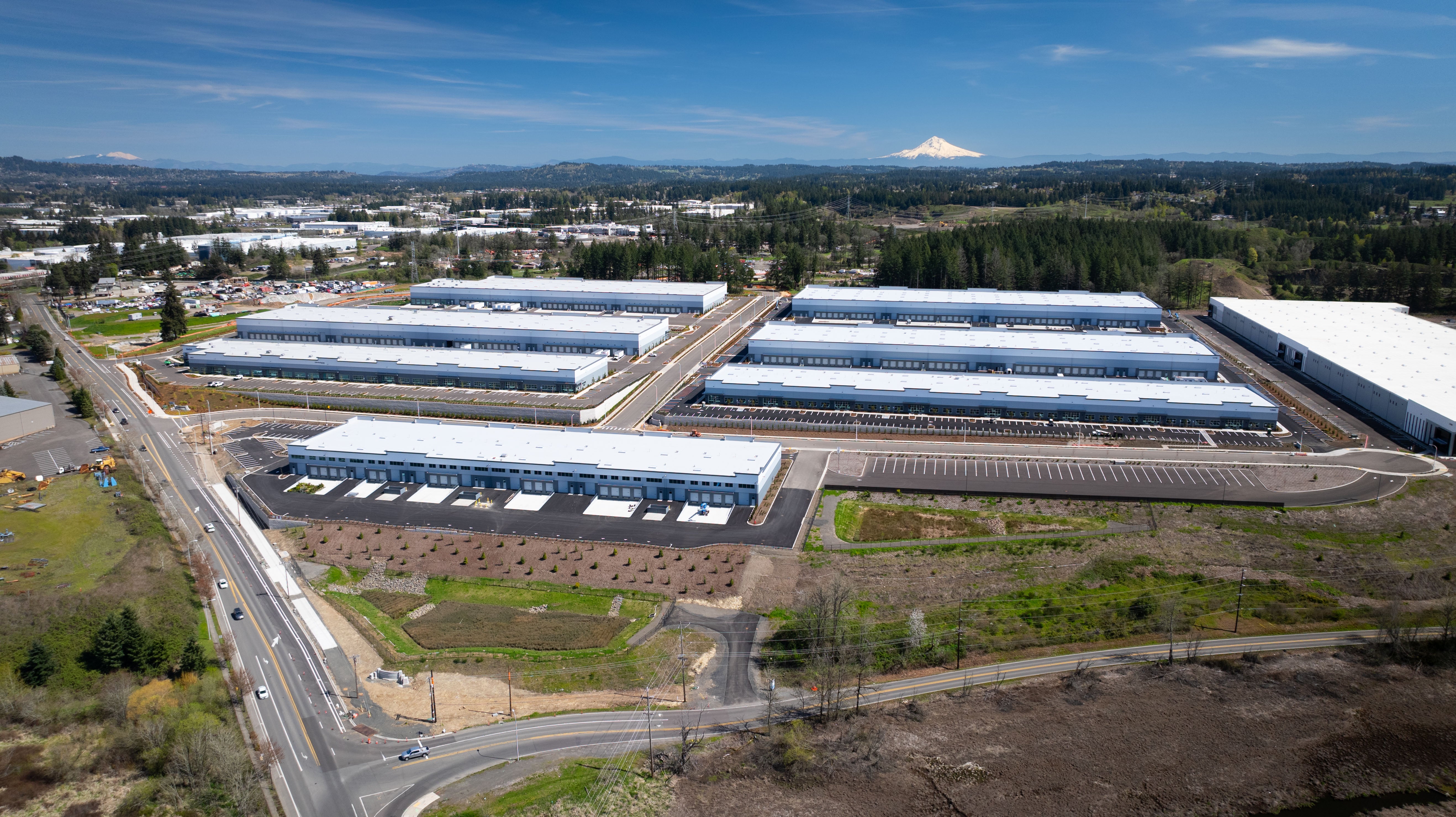 Aerial view of a large industrial complex with multiple warehouse buildings, parking lots, and nearby roads, surrounded by greenery and distant mountains under a blue sky.
