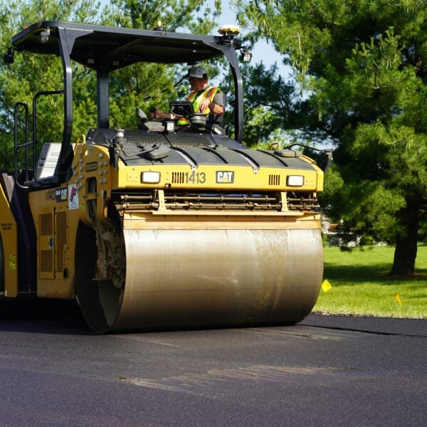 A construction worker operates a yellow road roller to smooth fresh asphalt on a sunny day near green trees.