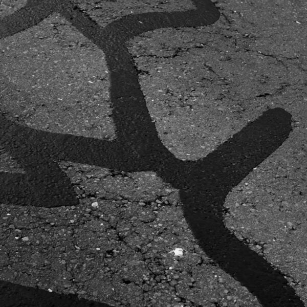 A close-up of cracked asphalt with irregular, dark lines of tar filling the gaps. The surface texture and patchwork pattern are visible.