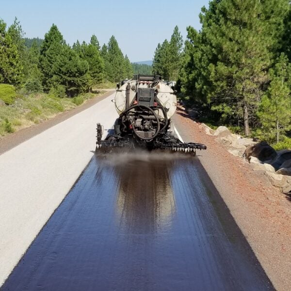 A road construction vehicle applies a layer of asphalt sealant to a rural road surrounded by trees and vegetation.