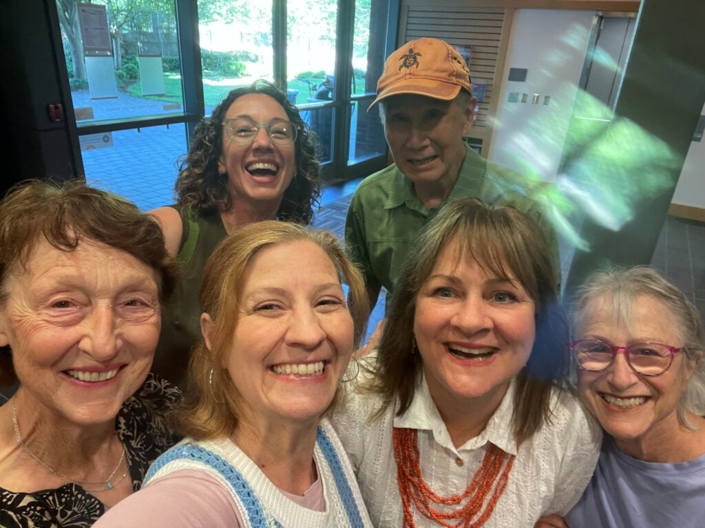 Six adults pose closely together indoors, smiling at the camera with windows and greenery visible in the background.