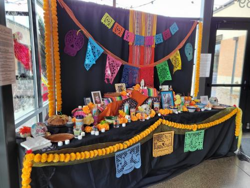 A Dia de los Muertos altar decorated with marigold garlands, colorful paper banners, framed photographs, and candles on a black tablecloth.