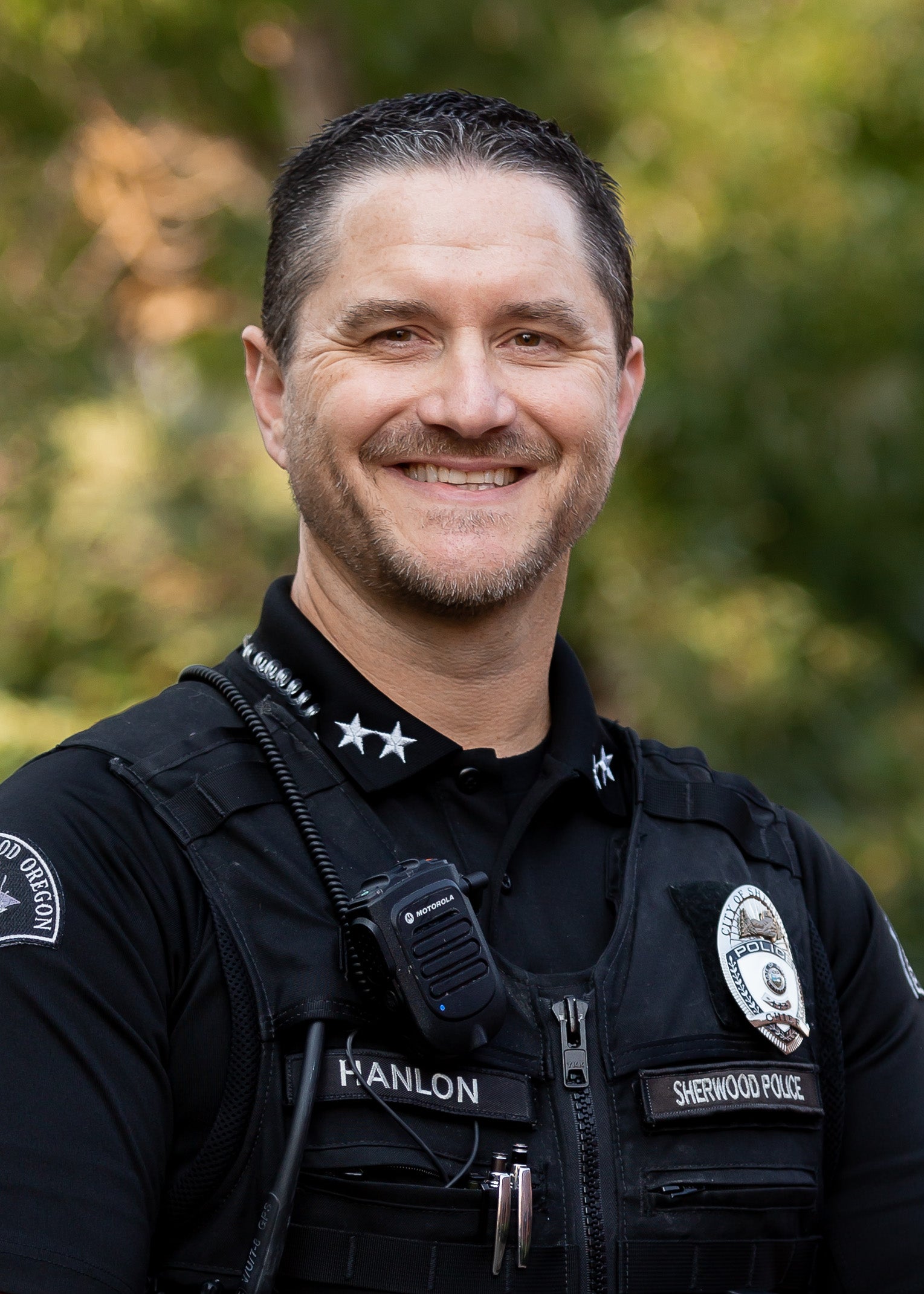 A smiling Sherwood Police officer in uniform with a badge, radio, and name tag.