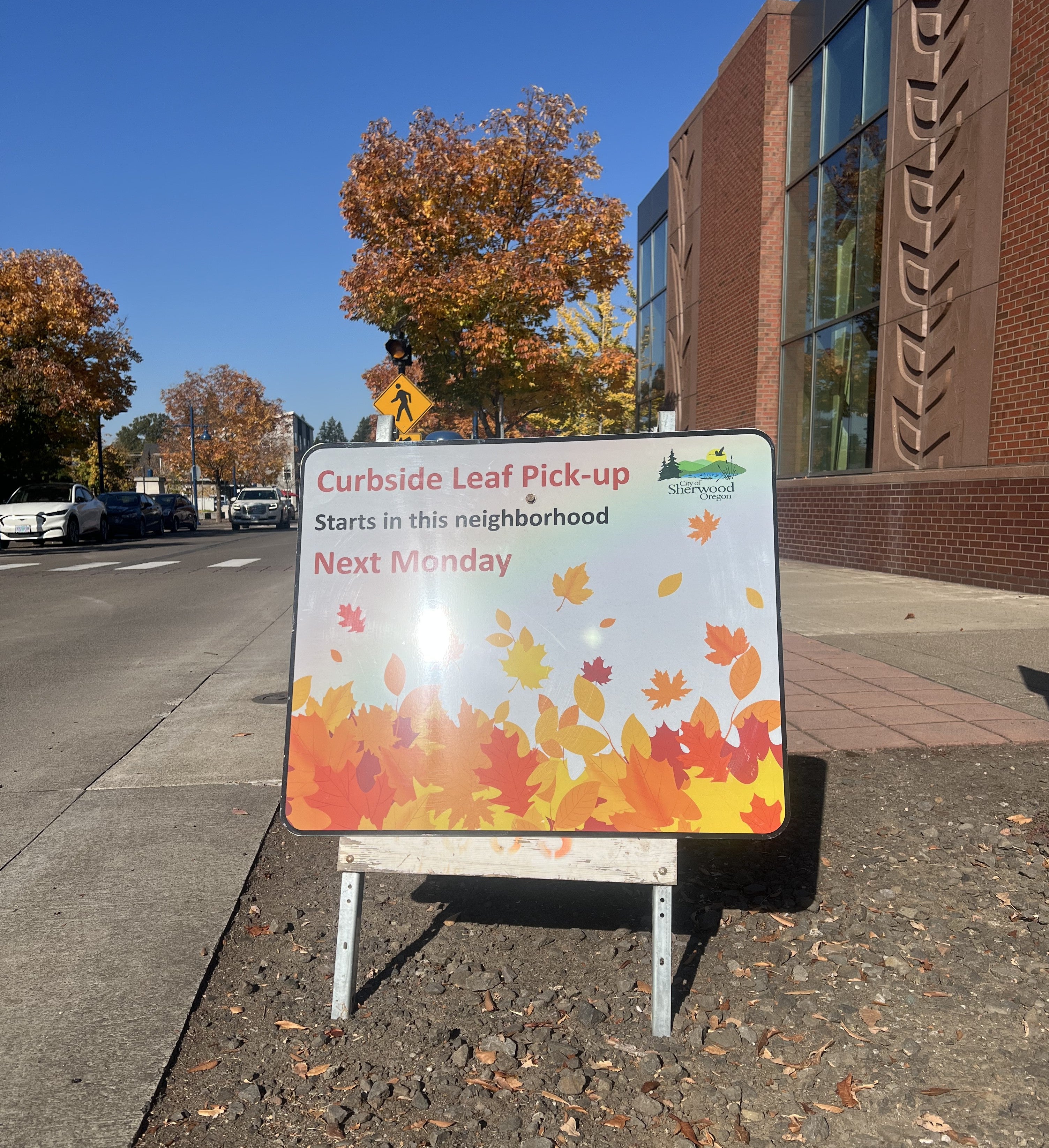 A sign on a sidewalk reads: “Curbside Leaf Pick-up Starts in this neighborhood Next Monday,” with the City of Sherwood, Oregon logo and a graphic of falling autumn leaves.