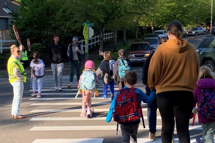 A group of children with backpacks and adults cross a street at a crosswalk on a sunny day, guided by a crossing guard in a yellow vest.