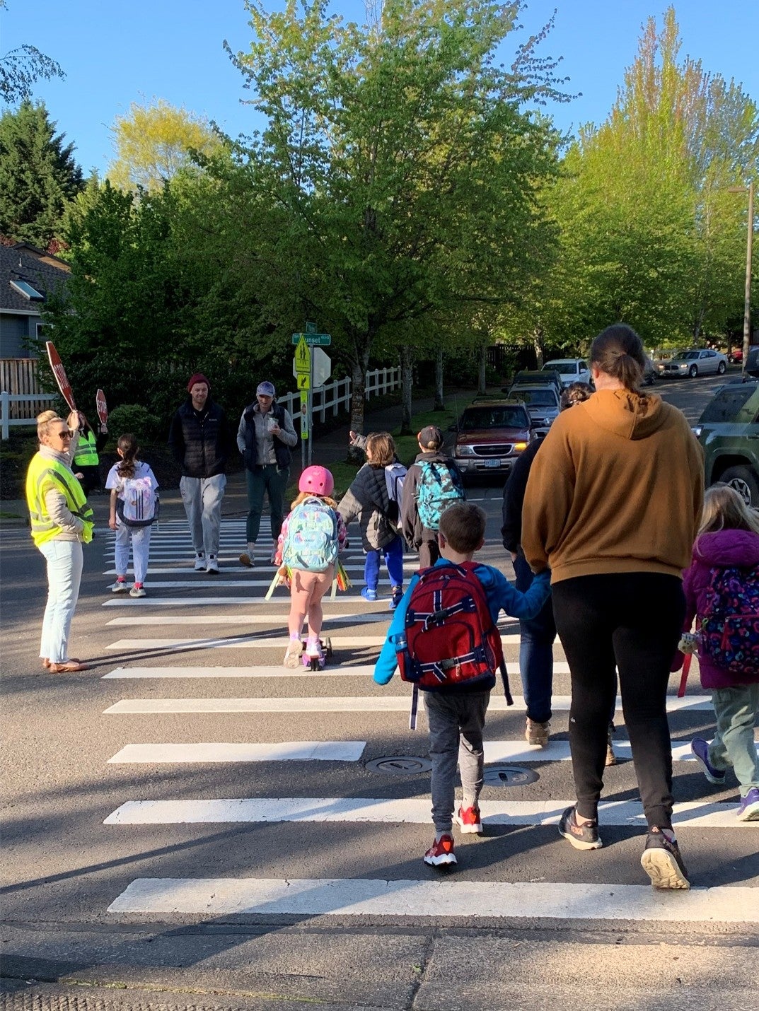 A group of children with backpacks and adults cross a street at a crosswalk on a sunny day, guided by a crossing guard in a yellow vest.